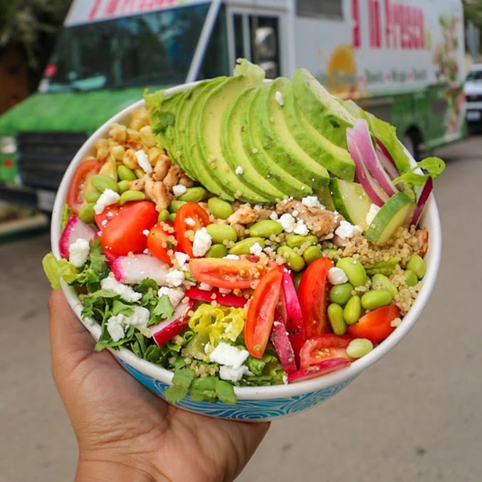 A Lo Fresco's healthy bowl with avocado, tomatoes, edamame, and feta cheese, held in front of a food truck.